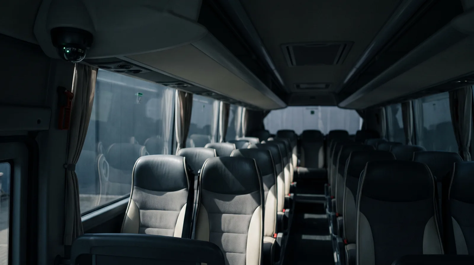 Interior de um ônibus rodoviário vazio ao entardecer, poltronas em fuga pelo corredor e uma pequena câmera dome fixada no teto junto ao para-brisa, com um reflexo verde na lente